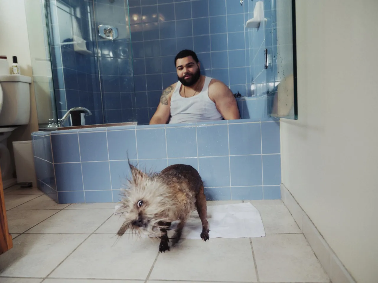 A man in a bathtub watches his wet dog shake off on the bathroom floor
